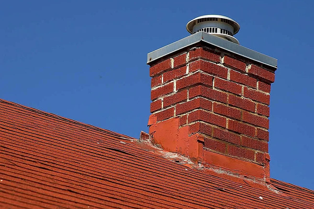 Red-brick chimney with a metal cap rising above a shingle roof against a blue sky